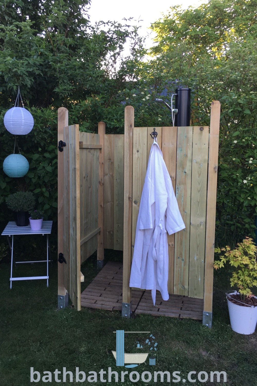 A rustic outdoor shower with towels hanging from its hooks, adjacent to a potted plant, creating a cozy and aesthetically appealing space. Perfect for outdoor shower ideas and garden inspirations, bringing charm to your backyard or pool area. For more unique ideas, visit bathbathrooms.com.