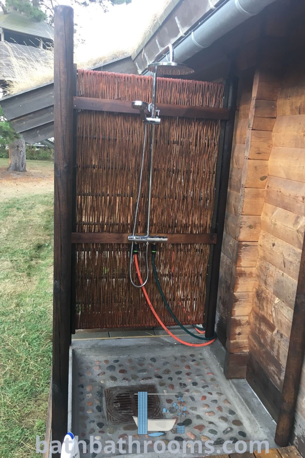 An outdoor shower in the corner of a wooden building with a hose, showcasing stylish eco bathroom features and outdoor shower ideas. This design combines functionality and aesthetics, ideal for unique decor inspirations. Discover more at bathbathrooms.com.