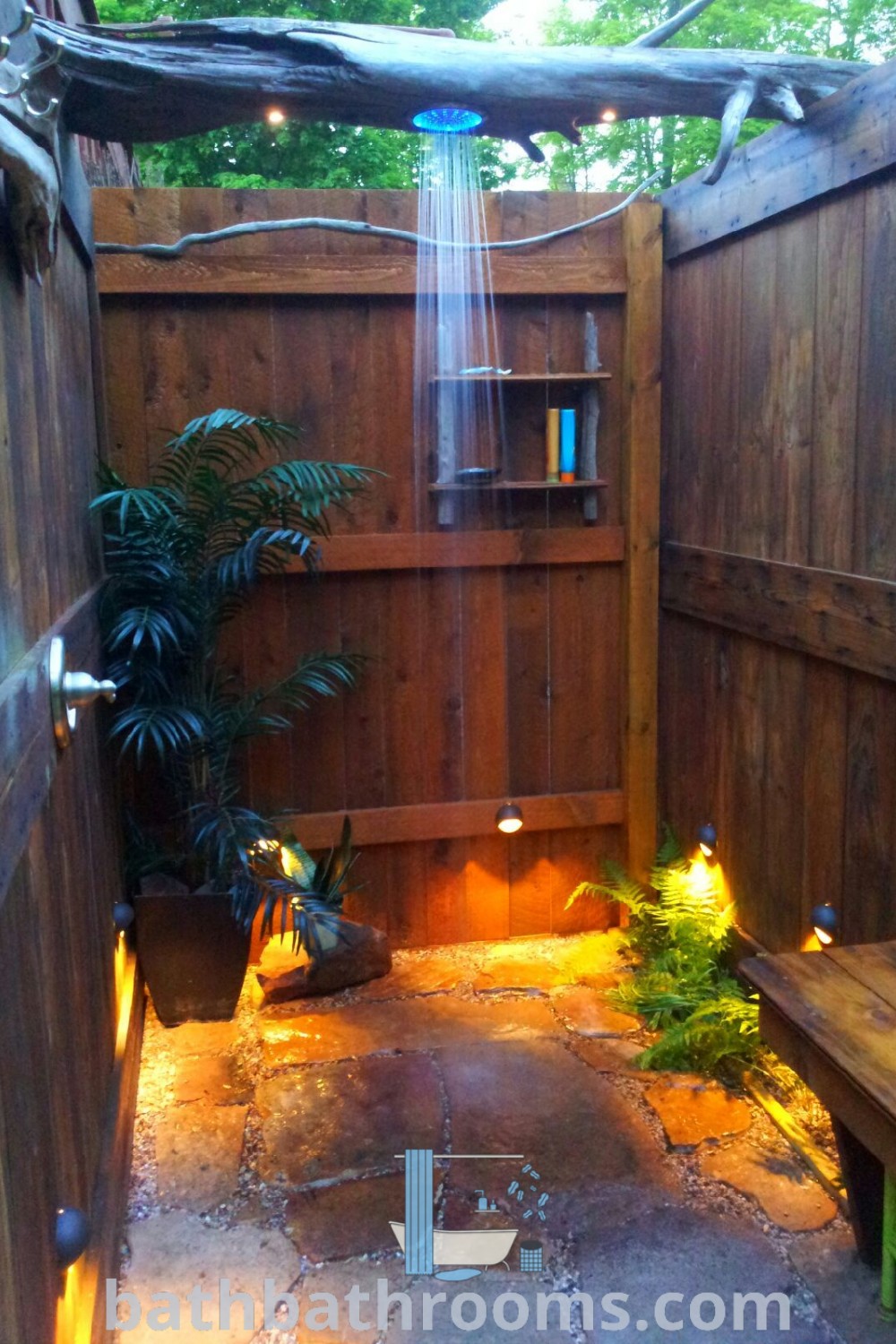 An outdoor shower with a concrete floor set in a wooden fenced area adorned with lights, showcasing rustic decor ideas and cozy aesthetic inspirations for backyard relaxation. Discover more about outdoor shower ideas at bathbathrooms.com.