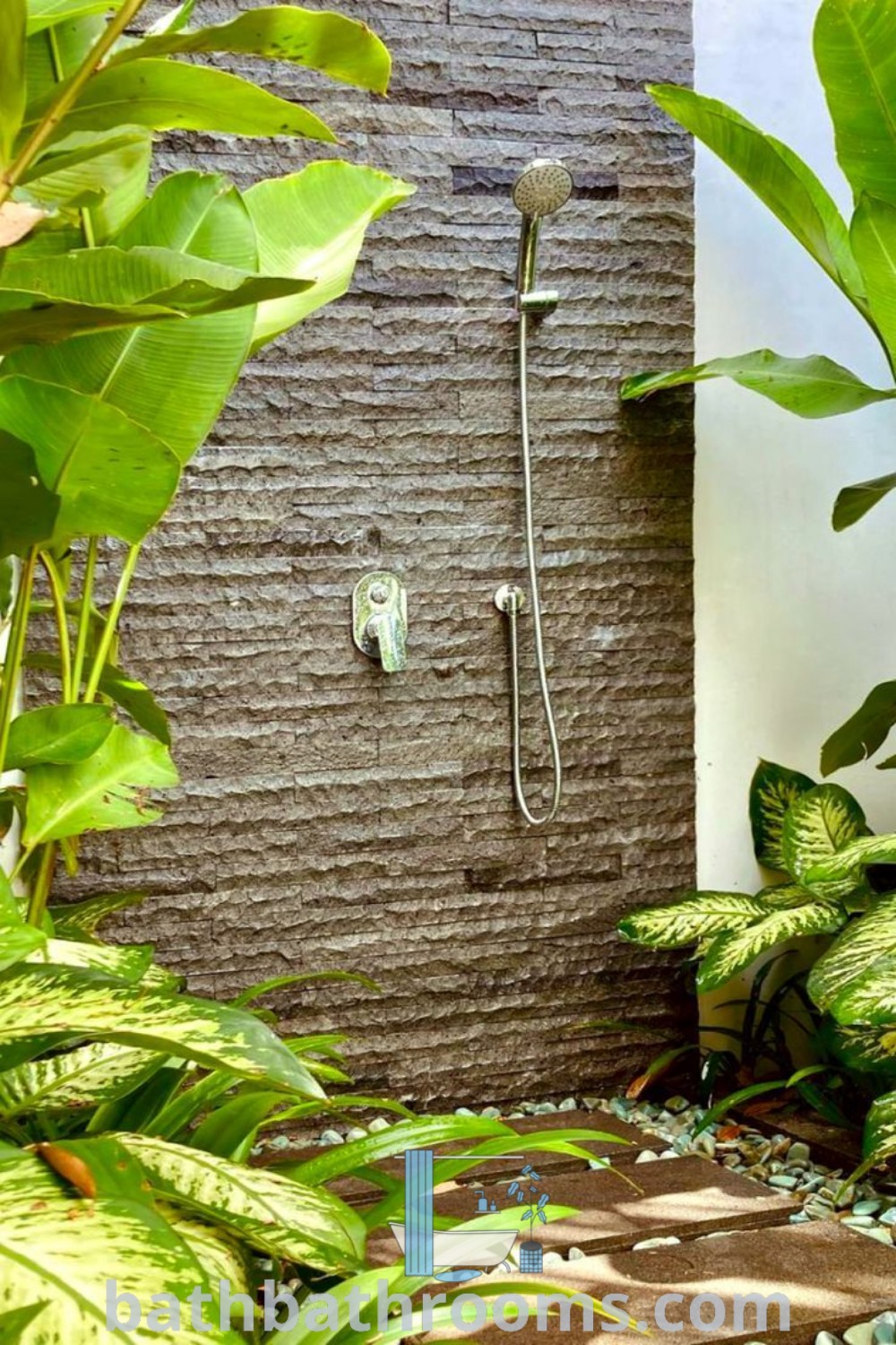 An outdoor shower surrounded by green plants, featuring gravel flooring and beautiful decor ideas for a cozy aesthetic. Discover unique outdoor shower designs and inspirations for your home at bathbathrooms.com.