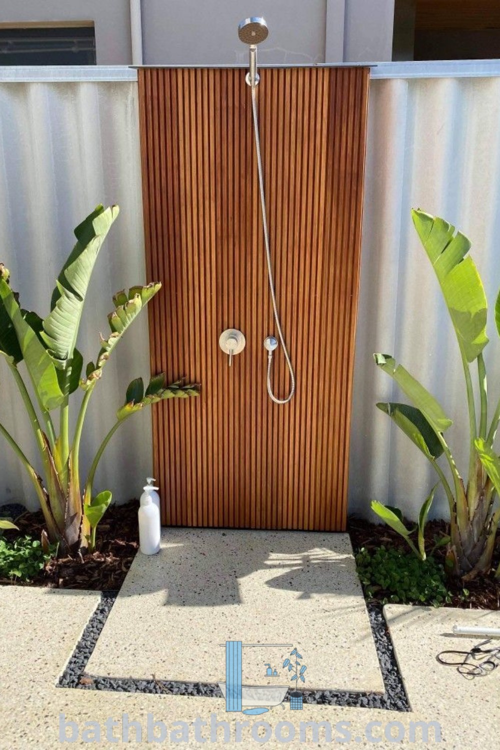 An outdoor shower is featured in front of a wooden door, surrounded by green plants, showcasing a blend of design ideas for outdoor showers and backyard pool landscaping. Explore more outdoor bathroom inspirations at bathbathrooms.com.