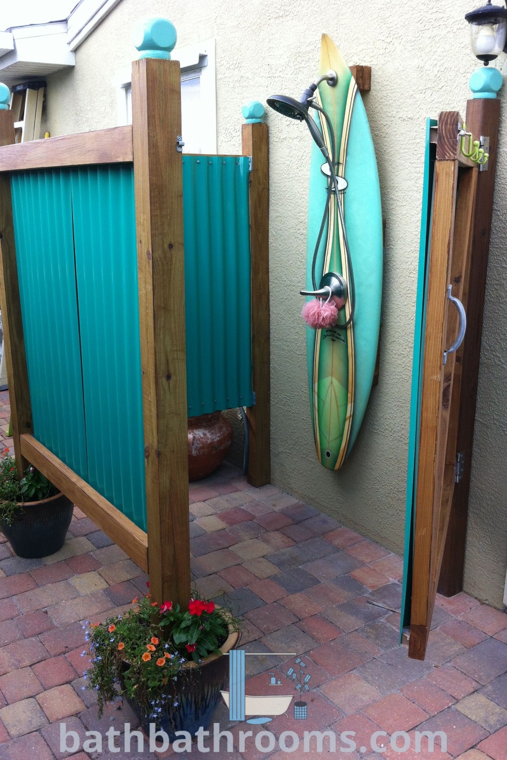 A surfboard mounted to the side of a wall adorned with flowers and potted plants, illustrating unique outdoor shower ideas that enhance your outdoor space. Discover inspiring design ideas for cozy outdoor showers at bathbathrooms.com.