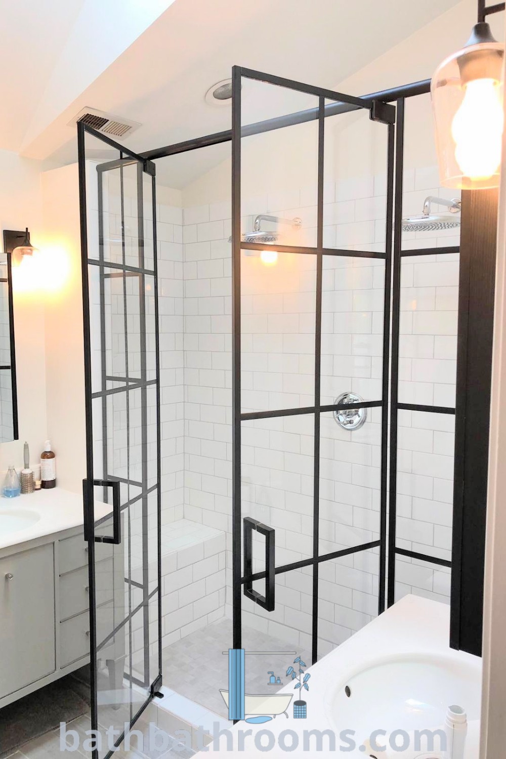 A beautiful bathroom showcasing a walk-in shower with black grid shower doors, a white sink, and a bathtub, all illuminated by two stylish lights. The decor features modern black shower hardware and embodies cozy design ideas for homes, making it an inspiring example of bathroom aesthetics. Visit bathbathrooms.com for more unique ideas.
