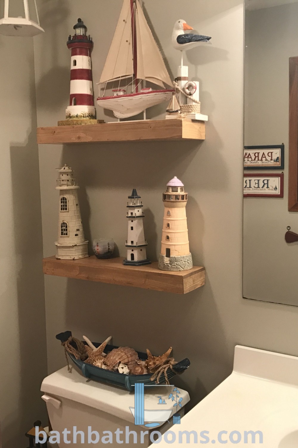 A cozy bathroom designed with a lighthouse theme, featuring two shelves above the toilet, a sink in front of a mirror, and nautical decor elements. Experience unique ideas and inspirations for your home at bathbathrooms.com.