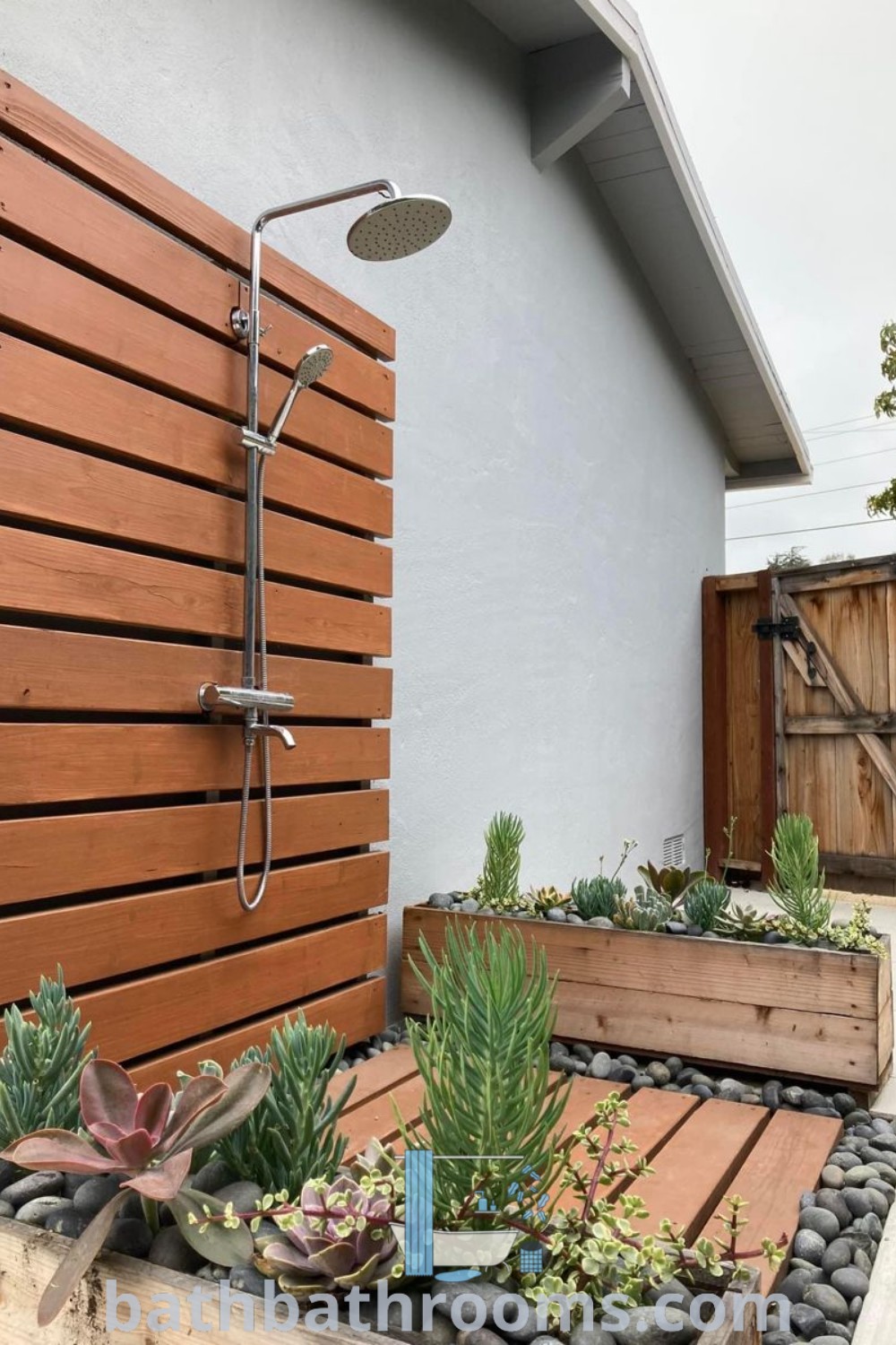An outdoor shower with wooden slats and succulents, set in a backyard with pavers, showcasing style and functionality. Explore outdoor shower ideas for your home at bathbathrooms.com.