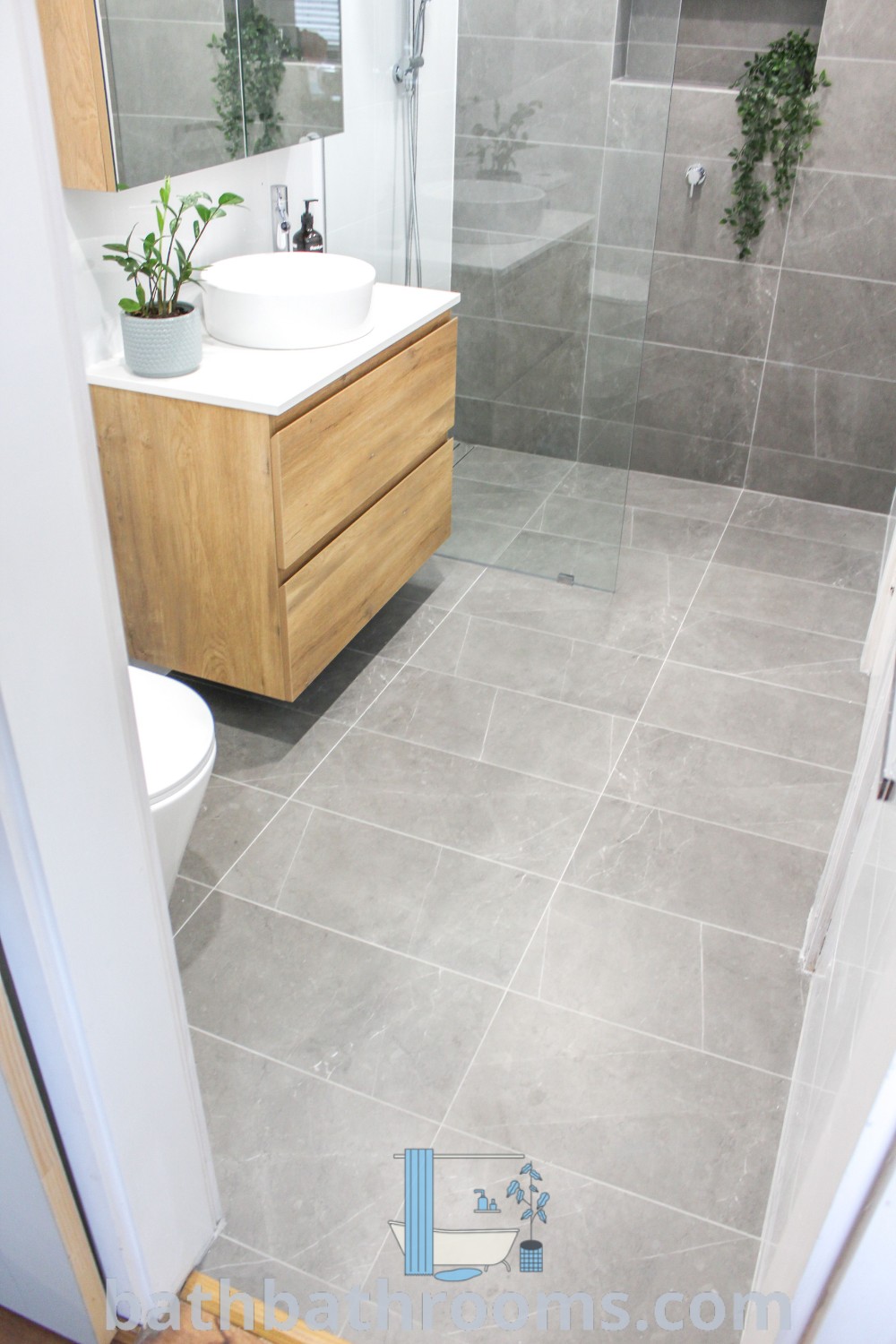 A small bathroom featuring grey tiles, a sink, toilet, and shower stall. This modern design reflects the cozy aesthetic with functional elements, showcasing inspiring ideas for small spaces. Discover more decor ideas at bathbathrooms.com.