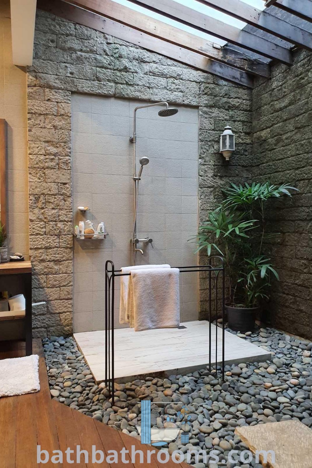 A rustic outdoor bathroom with stone walls and flooring, featuring two towels on a stand next to the shower, offering Zen house design and tropical bathroom ideas. Explore cozy decor inspirations for your home at bathbathrooms.com.