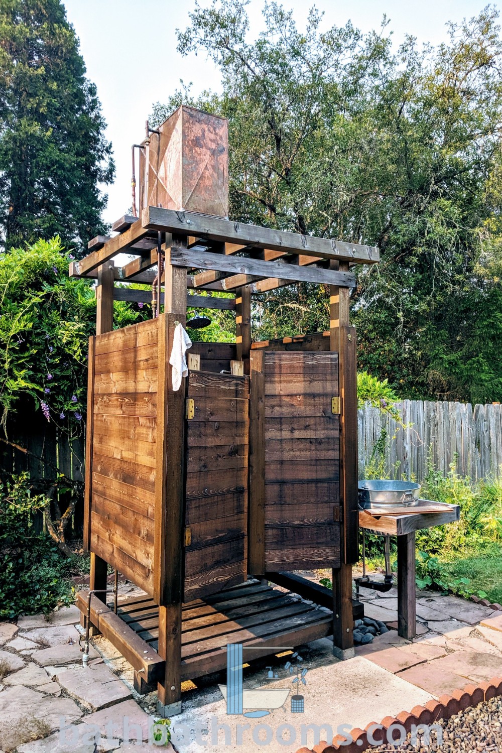 A wooden outhouse serving as an outdoor shower in a yard beside trees and a table, illustrated with cozy ideas for outdoor design. Explore patio yard ideas and unique outdoor shower inspirations at bathbathrooms.com.