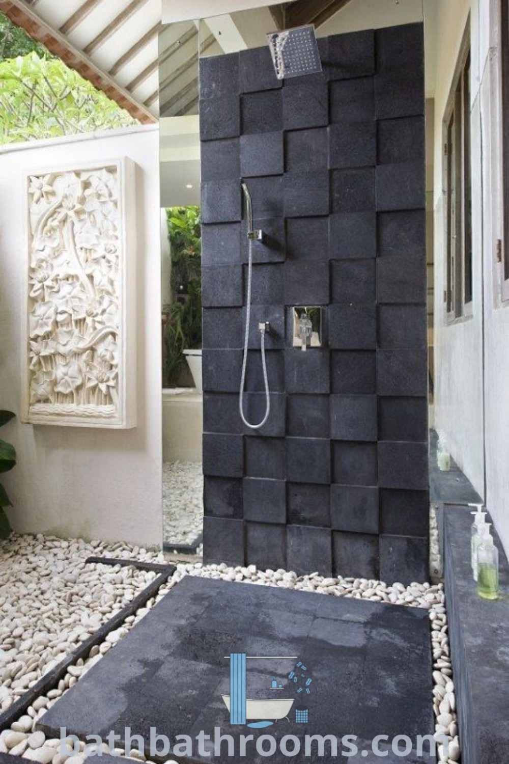 An elegant outdoor shower sits within a bathroom, next to a stone wall and floor, embodying inspiring outdoor bathroom design ideas. This setup highlights classic and balinese decor while maximizing style and space. Discover more cozy inspirations for your home at bathbathrooms.com.