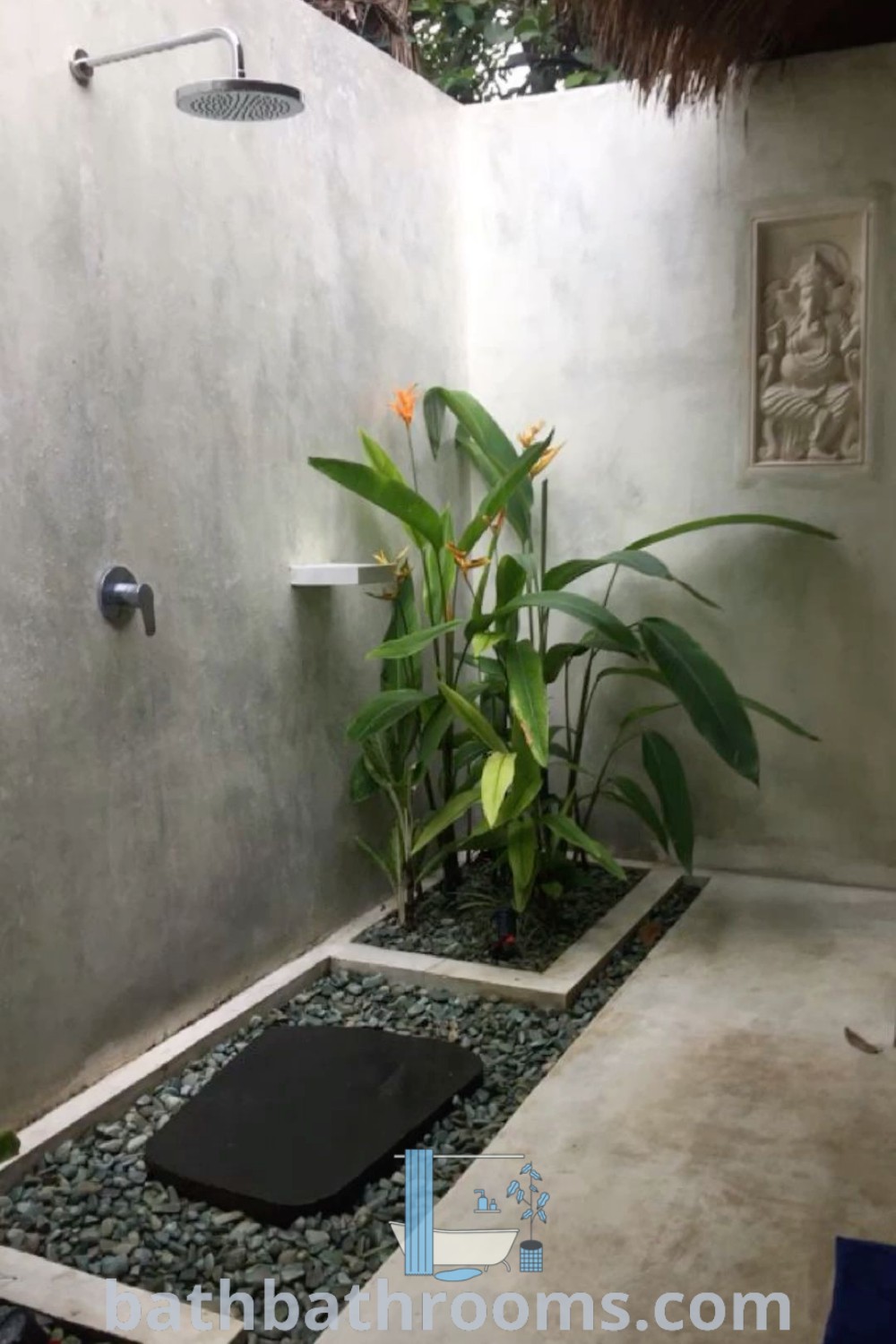 An outdoor shower surrounded by plants and rocks, featuring a cozy black mat on the ground. Explore outdoor bathroom ideas, designs, and inspirations for your home at bathbathrooms.com.