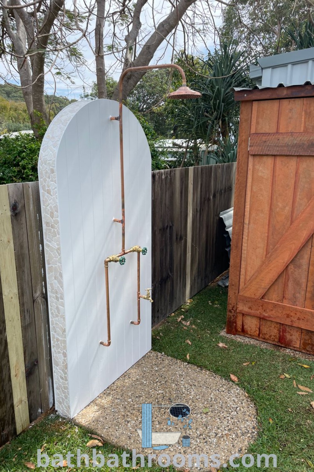 An outdoor shower in the middle of a yard, surrounded by a wooden fence and tree, designed to offer a stylish and functional space inspired by metro white decor ideas. This includes unique outdoor shower ideas to improve your backyard decor inspirations. Visit bathbathrooms.com for more design tips.