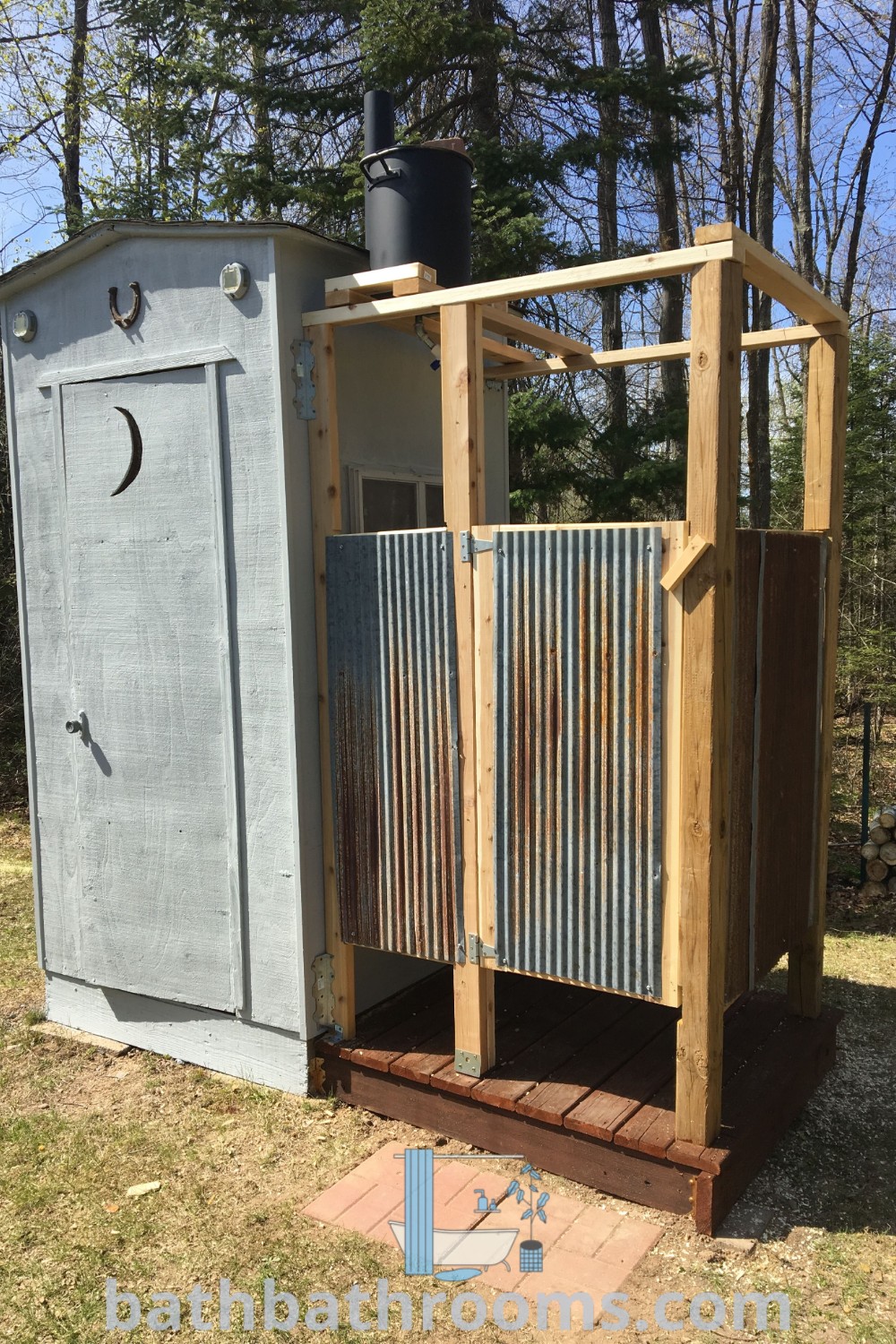 A cozy outhouse with a metal door surrounded by grass, featuring a shower combo. This outdoor toilet and shower idea showcases rustic charm and provides decor inspirations for a unique outdoor bathroom experience. Discover more cozy ideas at bathbathrooms.com.