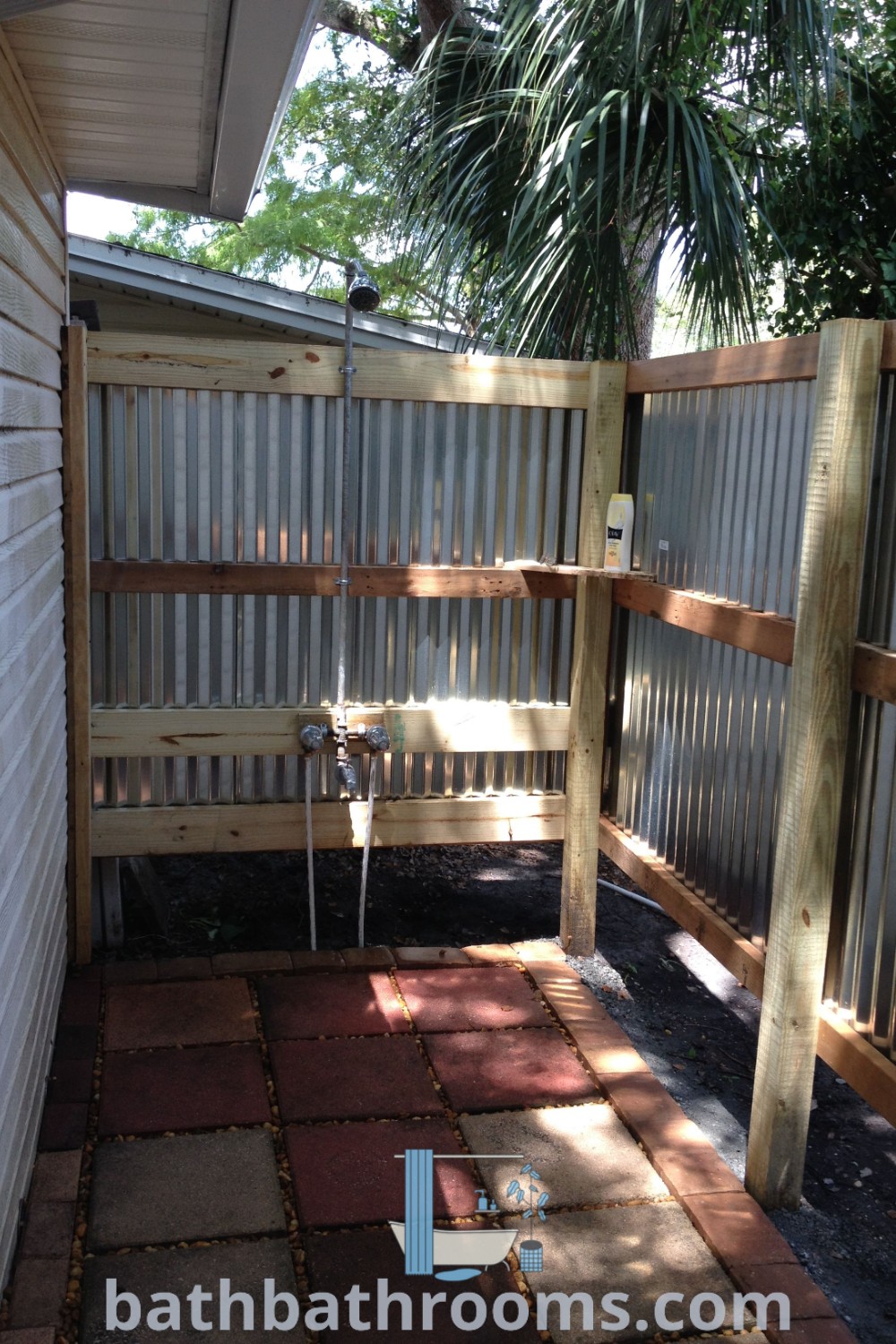 An outdoor shower situated in the middle of a yard, surrounded by brick pavers and a wood fence, exemplifying an outdoor bathhouse ideal for glamping aesthetics. This cozy setup offers inspiring ideas for shower enclosure designs and backyard decor. Discover more options at bathbathrooms.com.