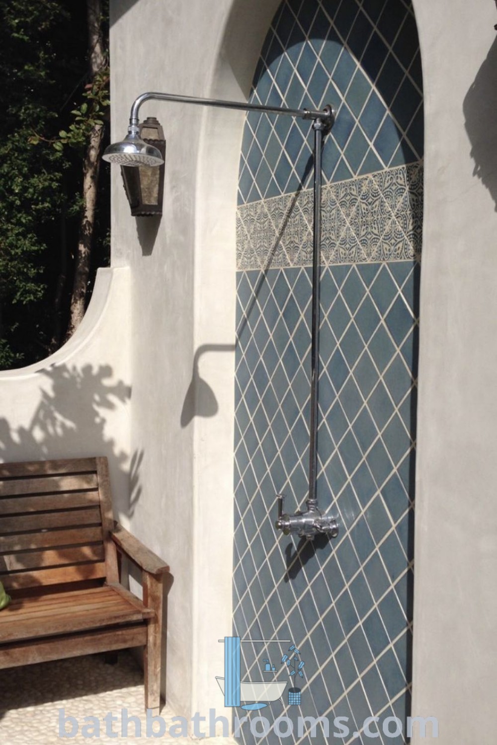 A wooden bench in front of a blue tiled shower stall, surrounded by greenery, epitomizing outdoor shower inspiration. The design showcases a Bali Villa pool aesthetic with Mediterranean boho elements, perfect for cozy home decor ideas. Discover more inspirations at bathbathrooms.com.