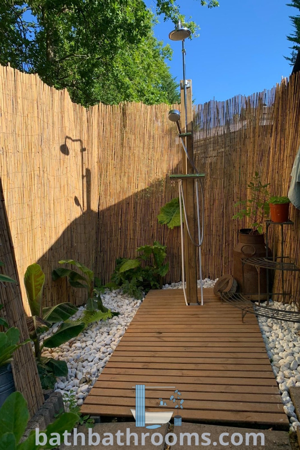 An outdoor shower surrounded by a wooden deck, lush plants, and rocks, showcasing cozy outdoor shower design ideas and rustic inspiration. Discover more design tips and decor ideas at bathbathrooms.com.