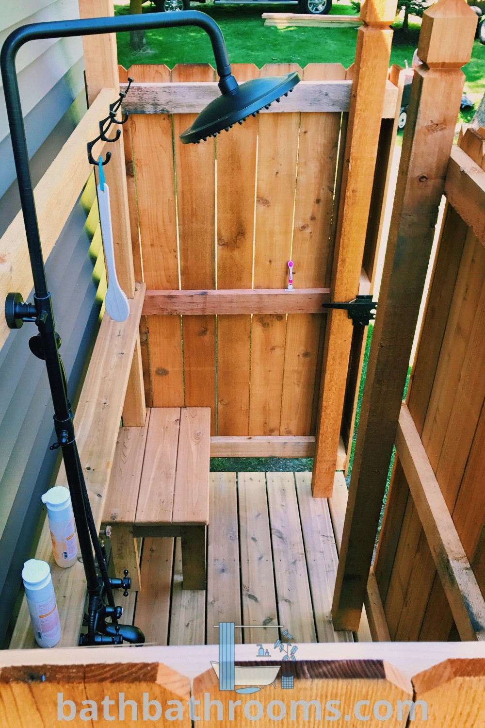 A cozy outdoor shower with a wooden fence and green grass in the background, complemented by a wood slatted privacy wall. This outdoor shower design inspires rustic aesthetics and functional beauty, perfect for cozy home decor ideas. Discover more on bathbathrooms.com.