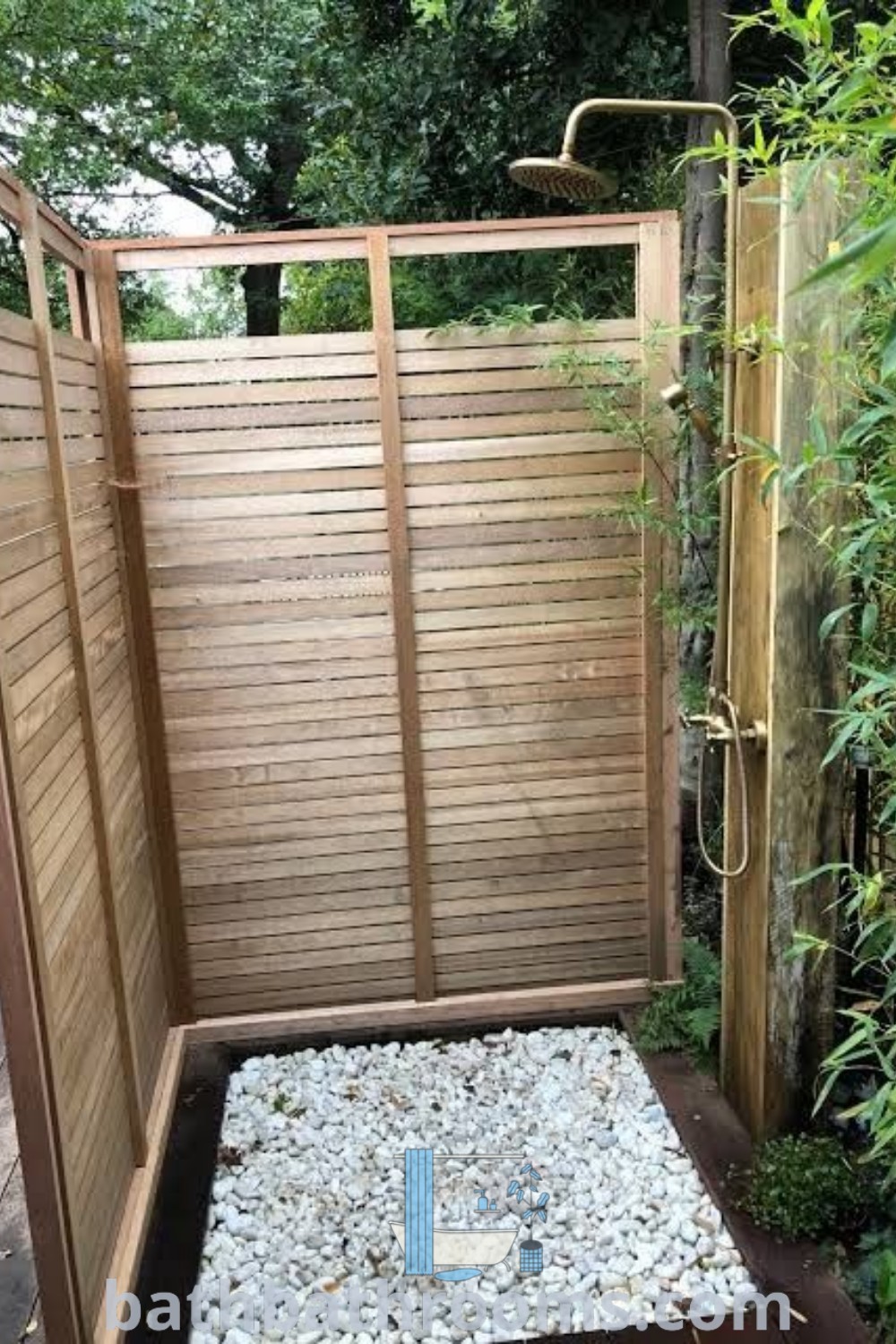 An outdoor shower with rocks and gravel next to a wooden fence, surrounded by greenery. This cozy design serves as an inspiring idea for backyard hardscape and outdoor toilet and shower setups. Discover more inspiring decor ideas at bathbathrooms.com.