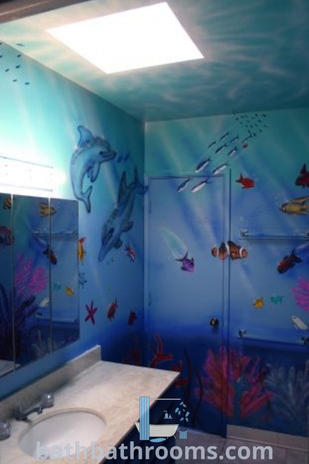A bathroom decorated in blue with dolphins and fish on the walls, featuring two sinks and an Aquariumcore 2000s style. The unique ocean-themed decor creates a vibrant and cozy aesthetic, making it an inspiring choice for anyone seeking aquatic bathroom design ideas. For more decor ideas, visit bathbathrooms.com.