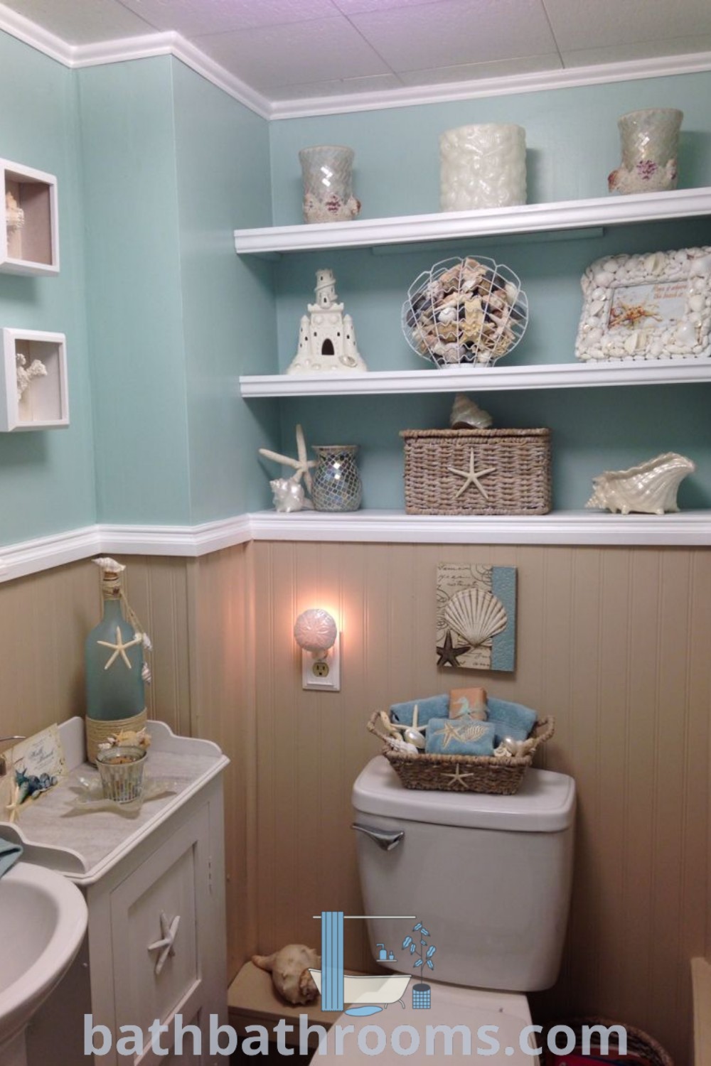 A beach-inspired bathroom decorated in blue and white, featuring shells on the shelf above the toilet. This cozy aesthetic exemplifies beach theme bathroom decor and coastal decor ideas, ideal for creating a tranquil atmosphere. Discover more inspirations for your home at bathbathrooms.com.
