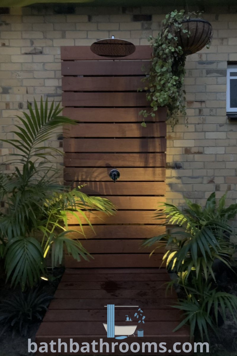 An outdoor shower surrounded by plants next to a brick wall and window, featuring cozy decor ideas and functional design for outdoor toilet and shower area. Explore outdoor shower ideas and inspirations at bathbathrooms.com.