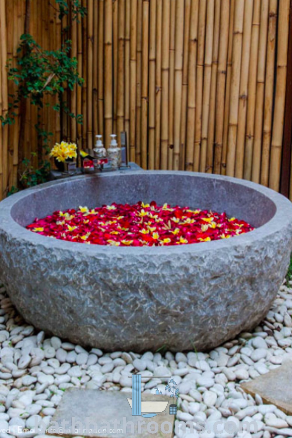 A large stone bathtub filled with flowers, nestled among rocks and gravel beside a bamboo fence, embodies a tranquil outdoor bathroom feel. Discover inspiring design ideas for your home on bathbathrooms.com.