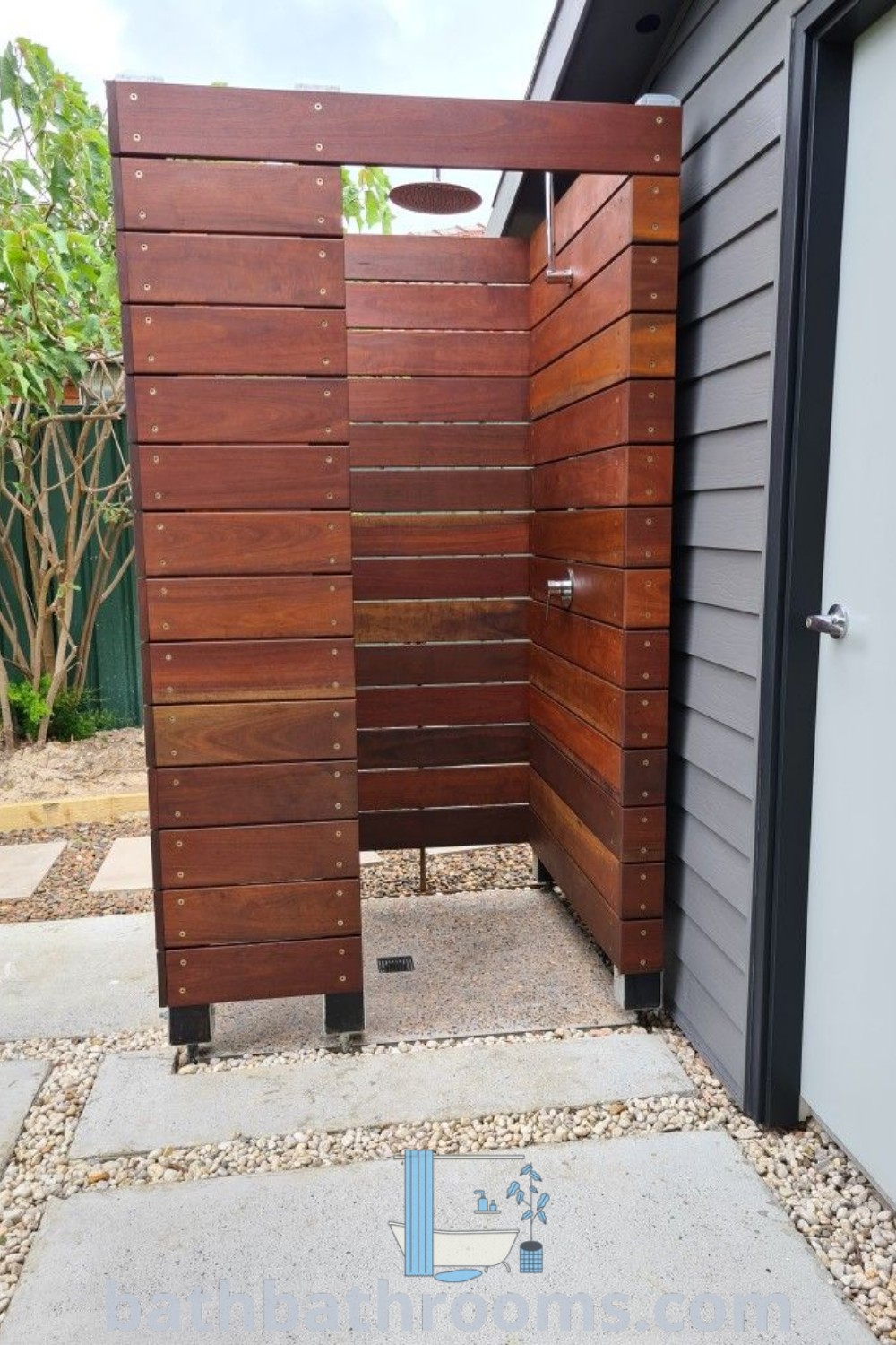 An outdoor shower next to an open wooden door, surrounded by trees and a brick walkway, exemplifying coastal outdoor shower ideas. Explore more about this inviting design and inspirations for your space at bathbathrooms.com.