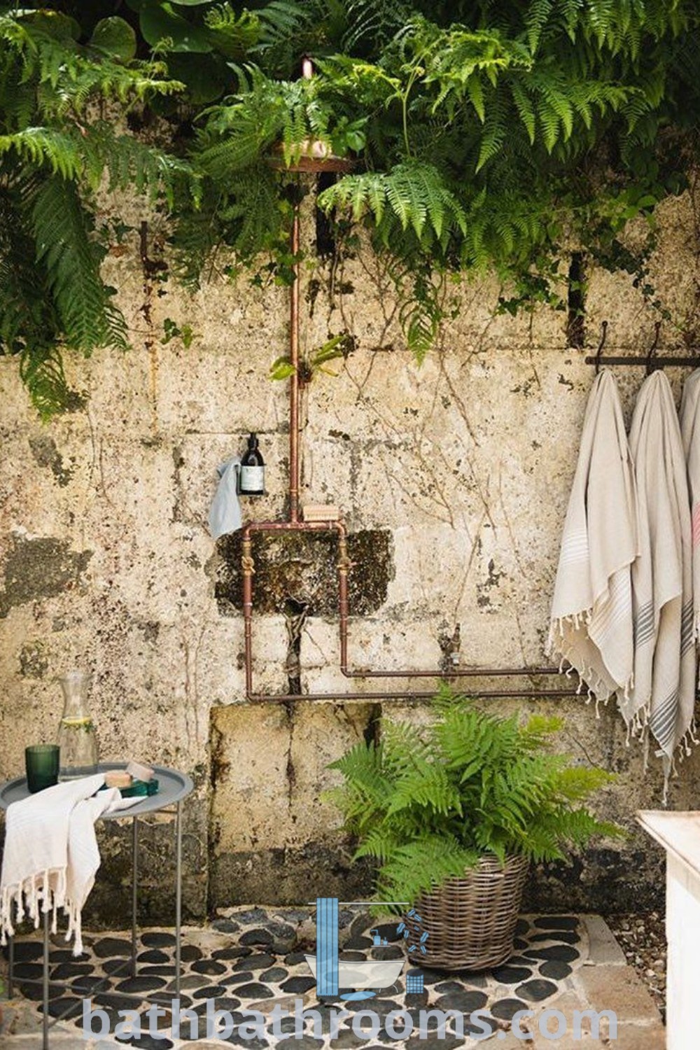 A rustic outdoor shower surrounded by plants and towels on its side wall, adjacent to a stone floor, illustrating an inspiring outdoor shower aesthetic for a cozy home. Perfect for outside bathrooms and patio designs. Discover more ideas at bathbathrooms.com.