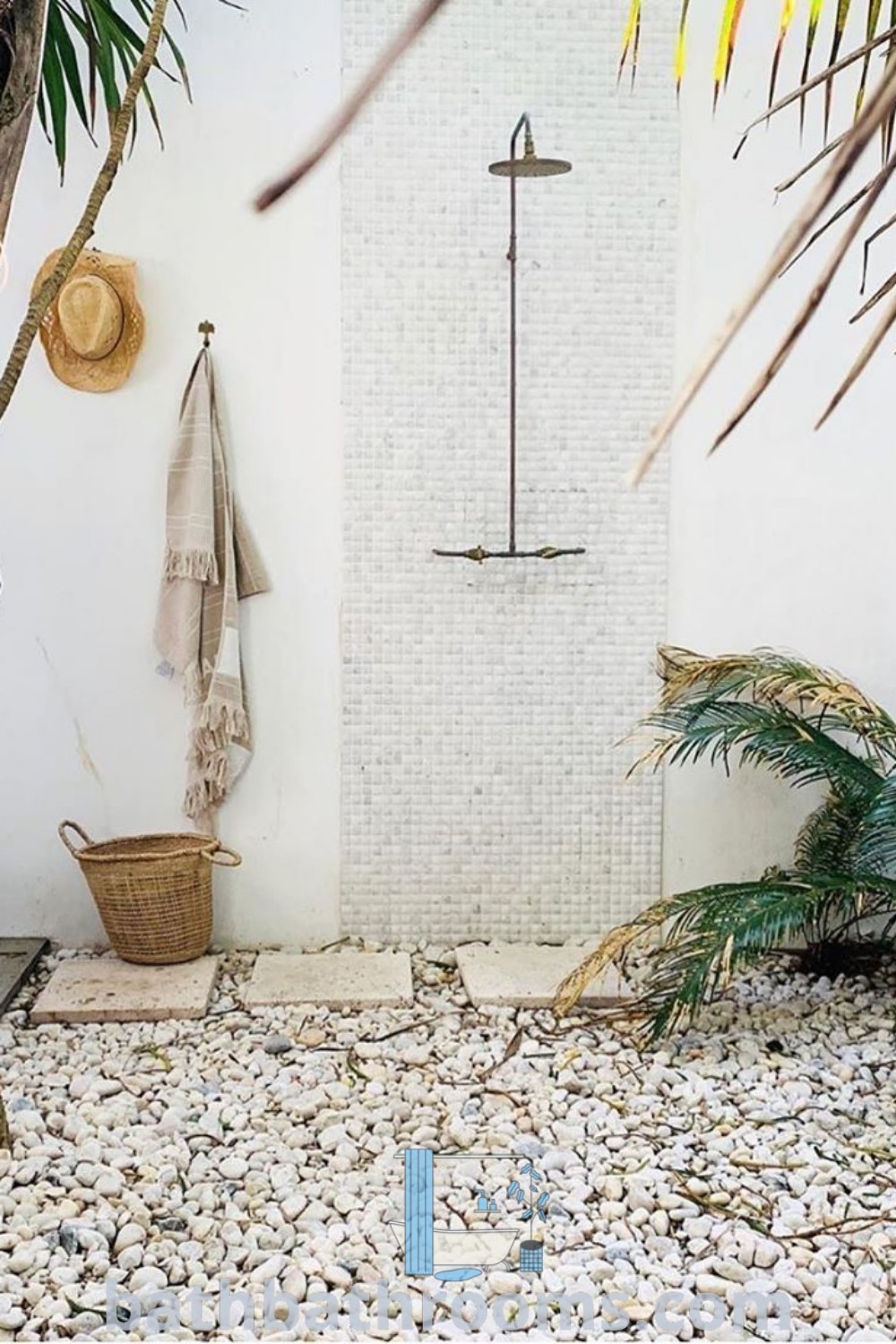 An outdoor shower surrounded by rocks and plants, next to a palm tree, showcasing outdoor shower landscaping ideas inspired by Ibiza and Bali. This cozy bath experience merges nature with functionality, featuring inspiring decor ideas for a unique outdoor setting. Discover more design inspirations at bathbathrooms.com.